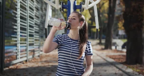 Жінка п'є воду з шейкера після тренування. Спортивна жінка п'є з фітнесу на деревах. Спортивна дівчина п'є з шакера. Дівчинка сидить на лавці, стомлена після тренування.. — стокове відео