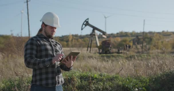Detailní záběr atraktivního inženýra dohlížejícího na čerpání ropy v blízkosti jednotky pomocí digitálního tabletu. Inženýr v klobouku používá počítač tabletu — Stock video
