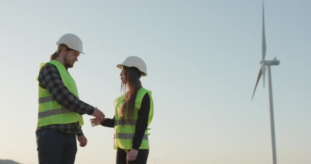 Inženýři muž a žena v helmách třesoucí se ruce na pozadí větrného mlýna turbíny. — Stock video
