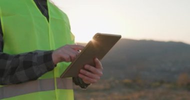 Kasktaki mühendis Rüzgar Türbinleri Enerji Santrali gün batımında elektronik tabletle fabrikada çalışıyor. Baş Mühendis Tablet Bilgisayar Kullanıyor.