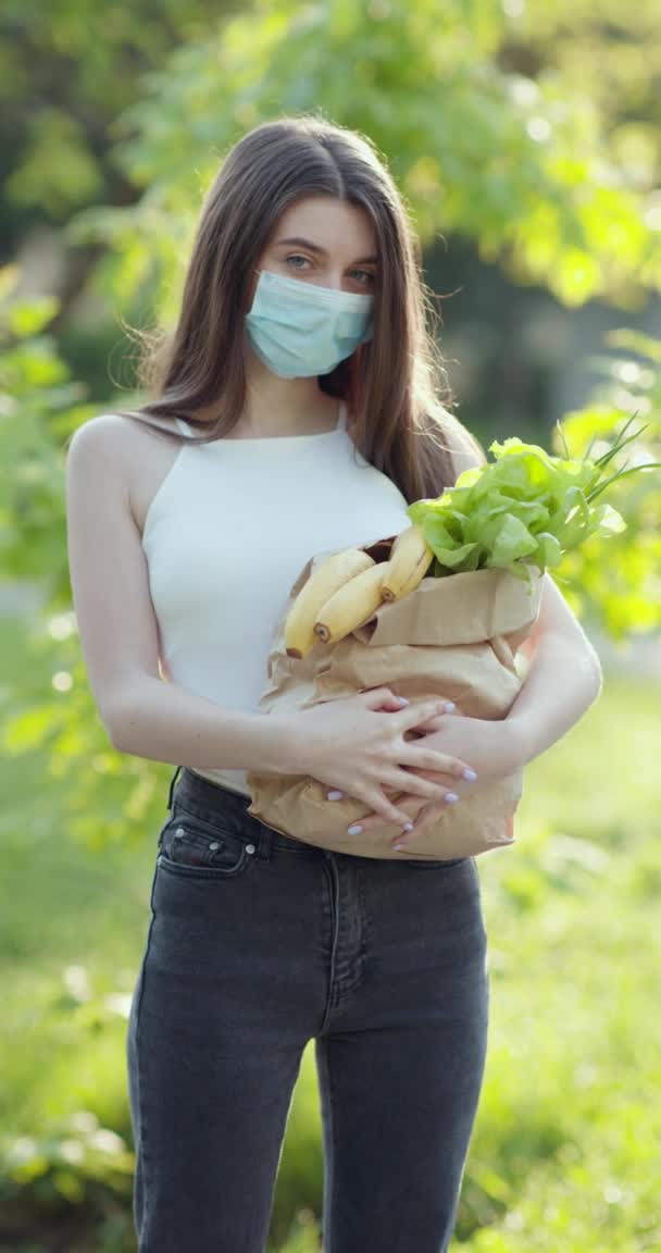 Vertikales Porträt einer jungen hübschen kaukasischen Zustellerin in medizinischer Maske im Park, die ein Päckchen mit frischen Lebensmitteln trägt. — Stockvideo