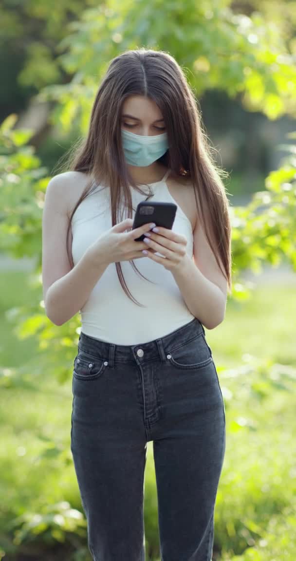 Vertical disparo joven mujer inteligente en una máscara médica está enviando mensajes de texto en el teléfono en el parque. Cuarentena COVID-19 en Europa. Transmisión del coronavirus en el parque público — Vídeos de Stock