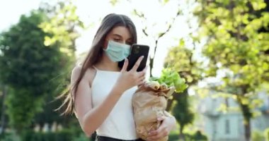Parkta akıllı telefon kullanan koruyucu maskeli genç bir kadın. Endişeli kız Covid-19 'la ilgili haberleri arıyor. Salgın Covid-19 koronavirüs koruması. Karantinaya yiyecek teslimatı.