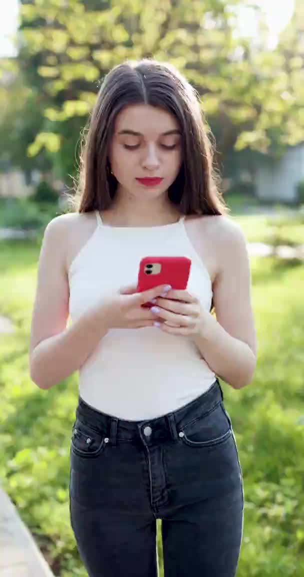 Vertical shot Woman uses phone texts scrolls surfs the internet search news. Young Woman Typing on Smartphone While Taking Walk in Park. — Stock Video