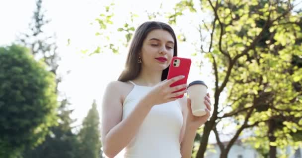 Menina atraente digitando mensagens em seu celular. Retrato de Pretty Woman Em pé no parque com Smartphone e xícara de café em suas mãos. Olhando enquanto digita Mensagens em sua conversa online — Vídeo de Stock