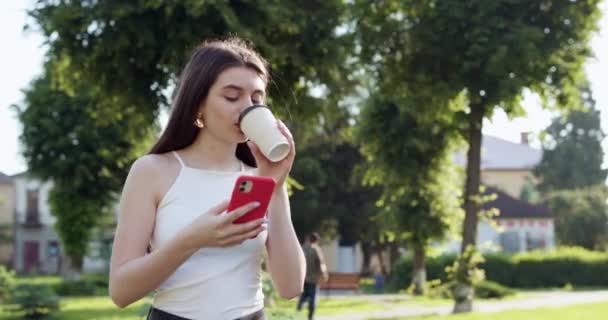 Sokak parşömenlerinde yürüyen ve modern akıllı telefonunda eğlenceli makaleler okuyan bir kız. Kupa 'yı sol elinde tutuyor. Güzel bayan telefonuyla meşgul.. — Stok video