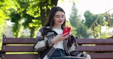 Moda kıyafetleri içinde güzel bir genç kadın şehir parkındaki bir bankta oturuyor ve aktif olarak telefonuyla mesajlar yolluyor. Çevrimiçi olmak, modern teknolojiler. Elinde sıcak bir içkiyle kağıt bardak tutuyor.