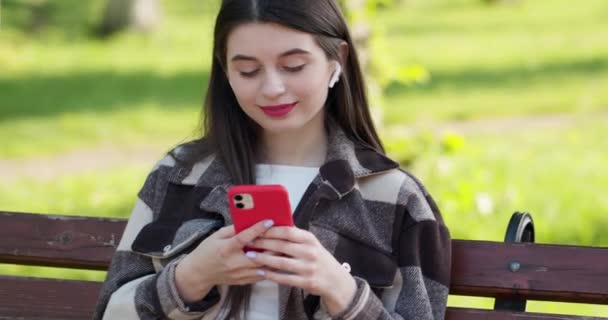 Meisje met koptelefoon en smartphone luistermuziek. Aantrekkelijke vrouw op de mobiele telefoon in het park. Stad, stedelijke achtergrond. — Stockvideo