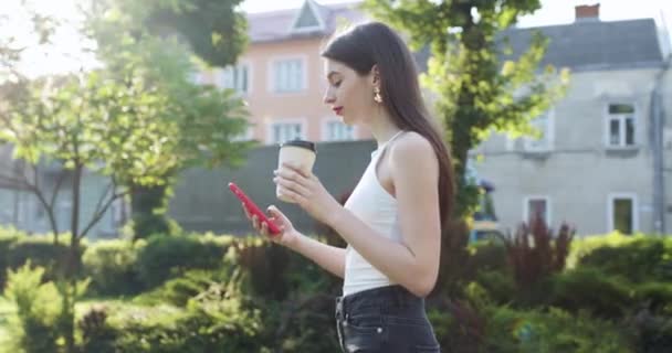 Dziewczyna śmieje się patrząc na swój smartfon. Krajobraz miejski z drzewami. Wesoła młoda dziewczyna z papierową filiżanką kawy w lewej ręce. Gorąca słoneczna pogoda. — Wideo stockowe