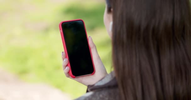 Schöne Mädchen hört Musik und verlässt durch das Band der sozialen Netzwerke auf ihrem Smartphone, während sie auf einer Bank im Park sitzt. — Stockvideo