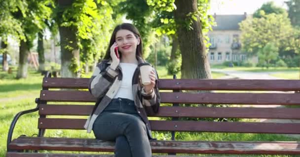 Prachtige Europese meisje maken van stem bericht over de telefoon. Jonge brunette zit op een bankje in een park. Mooie vrouw spreekt aan de telefoon en glimlacht — Stockvideo