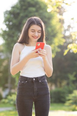 Kız gülüyor ve akıllı telefonundan bir şeyler okuyor. Parkta yürürken akıllı telefondan iletişim kuran neşeli genç bir kadın..