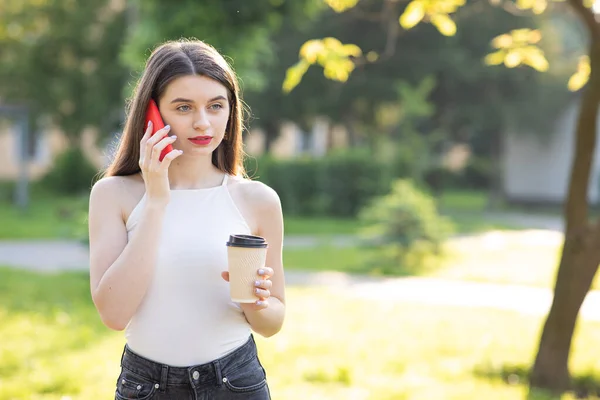 Ελκυστική κοπέλα δακτυλογραφεί μηνύματα στο κινητό της. Πορτρέτο της Pretty Woman Στέκεται στο πάρκο με Smartphone και καφέ φλιτζάνι στα χέρια της. Κοιτάζοντας ενώ πληκτρολογείτε μηνύματα στο διαδίκτυο Συνομιλία της — Φωτογραφία Αρχείου