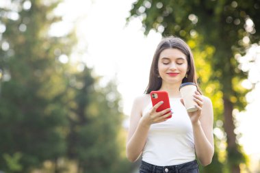 Akıllı telefonuna bakarken gülen bir kız. Ağaçlı şehir manzarası. Sol elinde bir fincan kahveyle neşeli bir genç kız.