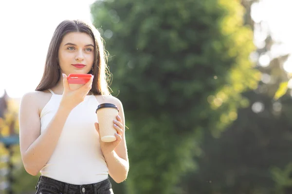 Dziewczyna chodząca po parku rozmawiająca przez smartfon. Happy girl za pomocą smartfona rozpoznawania głosu ai funkcji mowy głosowej na linii spacery w parku. Filiżanka z kawą w rękach — Zdjęcie stockowe