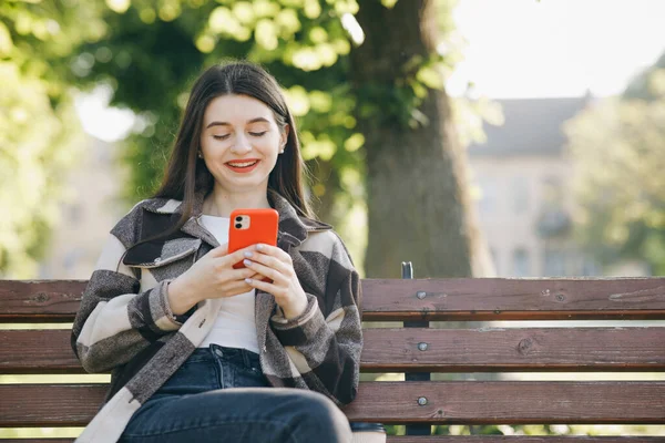 Молода жінка в модному повсякденному вбранні сидить на лавці в міському парку, активно пише повідомлення по телефону. Бути онлайн, сучасні технології . — стокове фото
