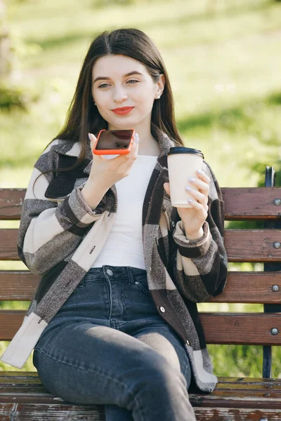 Κορίτσι που χρησιμοποιεί ένα έξυπνο τηλέφωνο φωνητικής αναγνώρισης ήχου ai λειτουργία ομιλίας μήνυμα. Γυναίκα με μοντέρνα casual στολή κάθεται σε ένα παγκάκι στο πάρκο της πόλης — Φωτογραφία Αρχείου