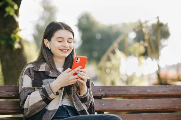 Dívka poslouchající hudbu v parku pomocí mobilního telefonu a nošení sluchátek. Atraktivní žena procházení na mobilním telefonu ve veřejném parku. Roztomilý mladý dívka v městském pozadí poslech hudby — Stock fotografie