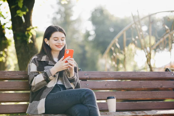 Portret dziewczyny noszącej słuchawki i używającej muzyki do słuchania smartfonów. Atrakcyjna kobieta przeglądająca telefon komórkowy w parku. Miasto, tło miejskie — Zdjęcie stockowe