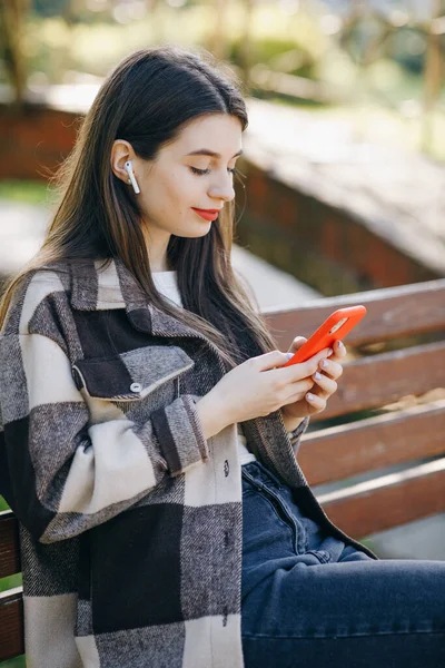 Fille heureuse écoutant de la musique dans le parc en utilisant un téléphone portable et en portant un casque. Attrayant femme naviguant sur le téléphone mobile dans le parc public. Jeune fille mignonne en fond urbain écoutant de la musique. — Photo