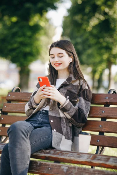 Pige iført øretelefoner og ved hjælp af smartphone lytter musik. Attraktiv kvinde browsing på mobiltelefon i parken. By, bybaggrund. - Stock-foto