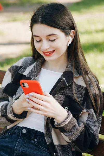 Jeune belle fille écoute de la musique et laisse à travers la bande des réseaux sociaux sur son smartphone tout en étant assis sur un banc dans le parc — Photo