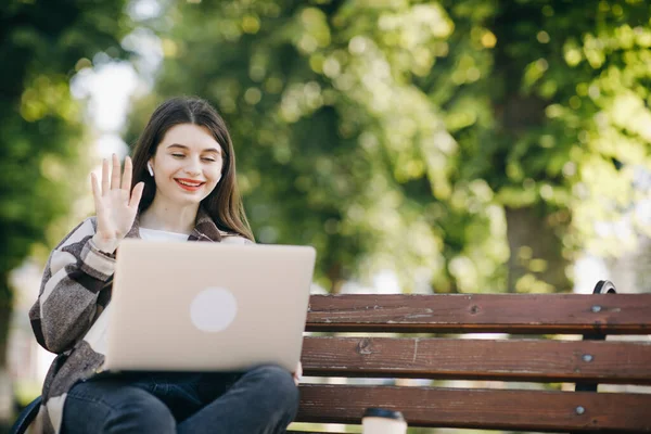 Girl is waving her hand to webcam on laptop while sitting on park bench. Student using webcam on laptop has video chat with friend. Caucasian woman smiling sitting on the bench lunch time outdoors. — Stock Photo, Image