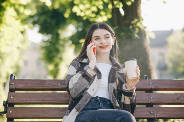 Młoda kobieta ubrana w stylowe ubrania, rozmawiająca przez telefon z chłopakiem o pomysłach na weekendy podczas przerwy w pracy, siedząca na ławce miejskiej z kawą na wynos. — Zdjęcie stockowe