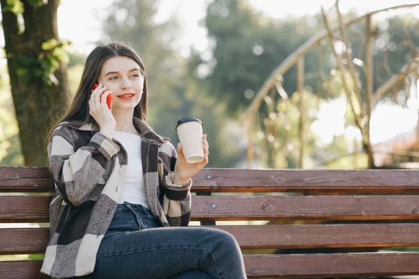 Młoda kobieta ubrana w stylowe ubrania, rozmawiająca przez telefon. Portret pięknej młodej dziewczyny rozmawiającej przez telefon, siedzącej na ławce w parku miejskim. — Zdjęcie stockowe