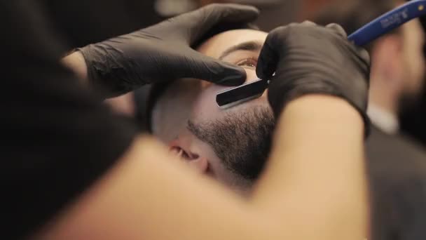 Профессиональный парикмахер бреет бороду клиента бритвой. Борода вырезана старомодным лезвием в парикмахерской. Красавчик сбривает бороду в парикмахерской. — стоковое видео