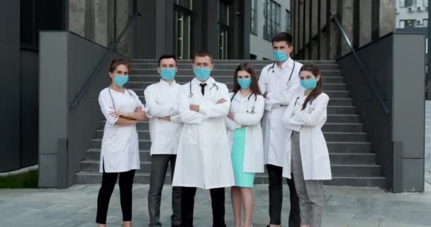 Los médicos se paran con los brazos cruzados mientras usan mascarillas protectoras al aire libre durante la epidemia de COVID-19 y mirando a la cámara — Vídeo de stock