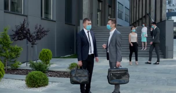 Zwei Mitarbeiter in Schutzmaske diskutieren vor dem Bürogebäude. Selbstbewusster Mann erklärt männlichen Kollegen etwas — Stockvideo
