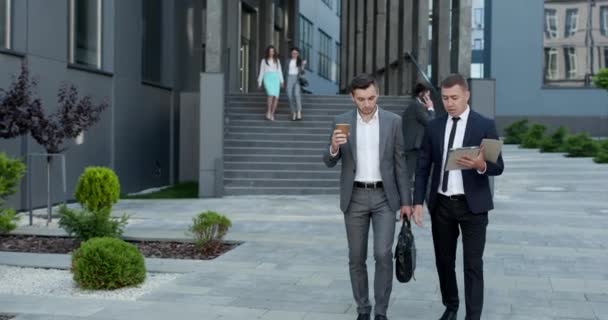 Dos hombres de negocios guapos con trajes y corbatas sosteniendo café en las manos y caminando desde el edificio de oficinas después del día de trabajo. Hombres elegantes hablando y discutiendo negocios. — Vídeos de Stock