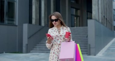 Günlük kıyafetli ve güneş gözlüklü genç bir kız alışveriş merkezinin önünde elinde çanta, telefon ve kağıt bardakla duruyor..