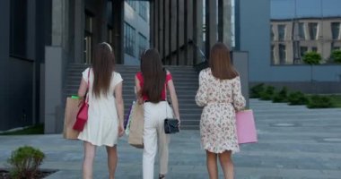 Ellerinde alışveriş torbalarıyla binaya doğru yürüyen, günlük kıyafetli üç genç kız arkadaş..