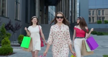 Alışveriş merkezinden ellerinde renkli poşetlerle çıkan bir grup mutlu genç kız. Güneşli Gün, Başarılı Alışveriş.