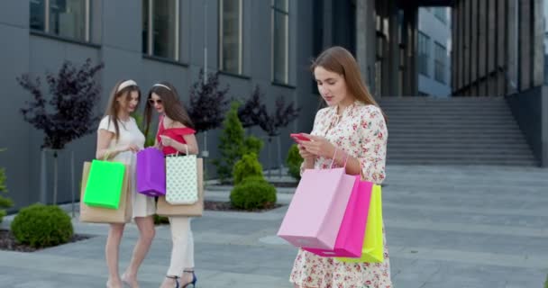 Молода красива дівчина стоїть перед торговим центром з телефоном і сумки в її руках. Група жінок на задньому плані . — стокове відео