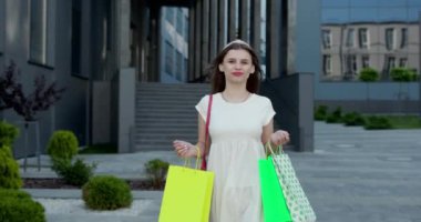 Beyaz elbiseli, kırmızı rujlu, alışveriş merkezinin önünde elinde çantalarla kameraya bakan genç ve güzel bir kız..