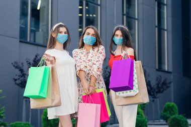 Giysileri, bluzu ve pantolonu olan kadınlar Coronavirus salgınını korumak için maske takıyorlar. Alışveriş merkezinin önünde ellerinde renkli poşetlerle duruyorlar..