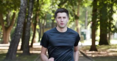 Spor üniformalı bir adam hız, mesafe ve kalp atışlarını ölçmek için şehir parkında akıllı adımlarla koşuyor. Kablosuz kulaklıklar. Dışarıda antrenman var. 4K HAM