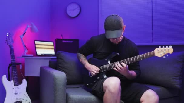 Ο κιθαρίστας Virtuoso παίζει ηλεκτρική κιθάρα στον καναπέ με χρωματιστά φώτα LED στο Home Studio. Ο άνθρωπος παίζει ηλεκτρική κιθάρα. Μουσικά βίντεο πανκ, heavy metal ή rock — Αρχείο Βίντεο