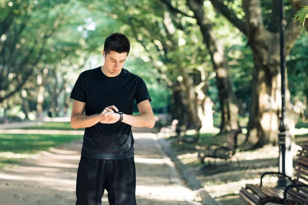 Aplicación a distancia. Hombre deportivo corriendo por el camino vacío en el parque comprobando la notificación de reloj inteligente en línea usando auriculares inalámbricos mientras entrenaba afuera. Vida activa saludable. — Foto de Stock