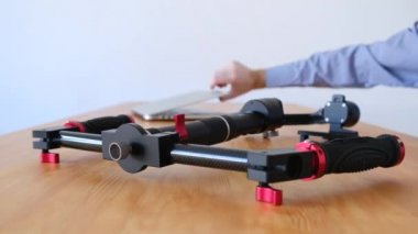 Mavi gömlekli kimliği belirsiz adam üç eksenli sağlamlık için video kamera ofis ahşap masa yatan için talimatları okumak için bir dizüstü bilgisayar açar. Profesyonel video üretim ekipmanları kavramı