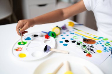 Child painting with colorful paints and tools on white table. High quality.