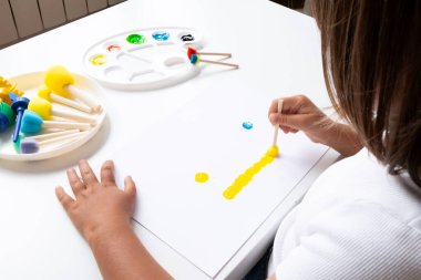 Young child painting with bright colors using sponges and sticks on white paper at home. High quality.