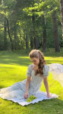 Uzun kıvırcık saçlı güzel bir kadın elinde şemsiye ve sepetle yeşil çayırlarda poz veriyor.