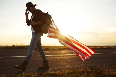 4 Temmuz. Amerikan Flag.Patriotic tatil. Amerika bayrağı ile seyahat. Adam bir şapka, bir sırt çantası, bir tişört ve kot pantolon giyiyor. Güzel gün batımı ışık. Amerikan tarzı. 