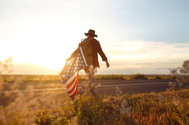 4 Temmuz. Amerikan Flag.Patriotic tatil. Amerika bayrağı ile seyahat. Adam bir şapka, bir sırt çantası, bir tişört ve kot pantolon giyiyor. Güzel gün batımı ışık. Amerikan tarzı. 