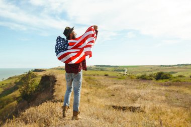 4 Temmuz. 4 Temmuz. Ulusal bayrağı olan Amerikalı. Amerikan bayrağı. Bağımsızlık günü. Vatanseverlik tatili. Adam şapka, sırt çantası, gömlek ve kot giyiyor. Güzel günbatımı ışık. 