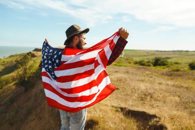 4 Temmuz. 4 Temmuz. Ulusal bayrağı olan Amerikalı. Amerikan bayrağı. Bağımsızlık günü. Vatanseverlik tatili. Adam şapka, sırt çantası, gömlek ve kot giyiyor. Güzel günbatımı ışık. 